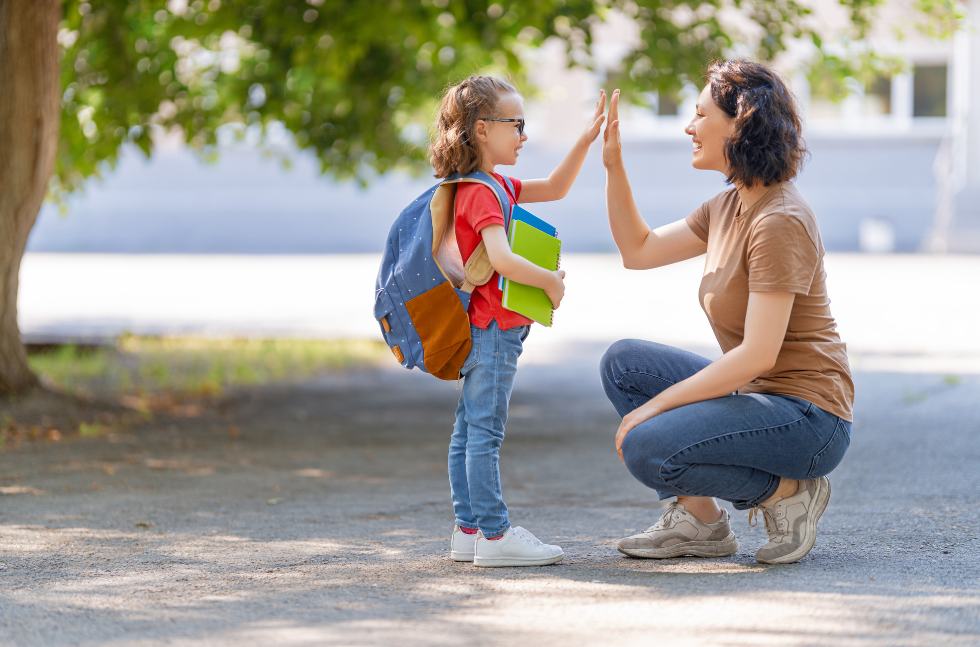 Beduerfnisorientierte-Erziehung-Paedagogik-Lehrerin-freundlich-klatsch-schuelerin-ab Frau in der Hocke und Mädchen im Grundschulalter mit geschulterter Schultasche klatschen sich ab